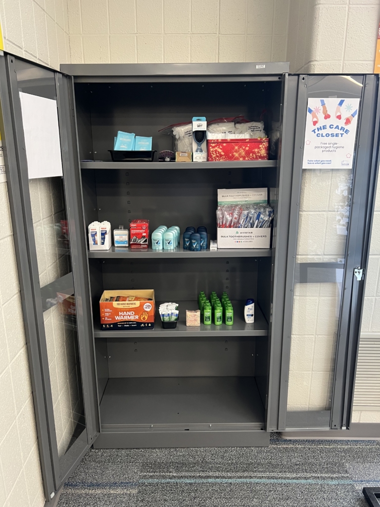 Image of the Taylor Library Care Closet. A tall, metal shelving unit with doors open wide. There are 3 shelves filled with various items. On the top shelf: body wipes, hairbrushes, & pads. On the second shelf: bandages, cotton swabs, deodorant, toothpaste, & toothbrushes. On the third shelf: hand warmers, petroleum jelly, soap, & shampoo. The bottom of the shelving unit is empty.
